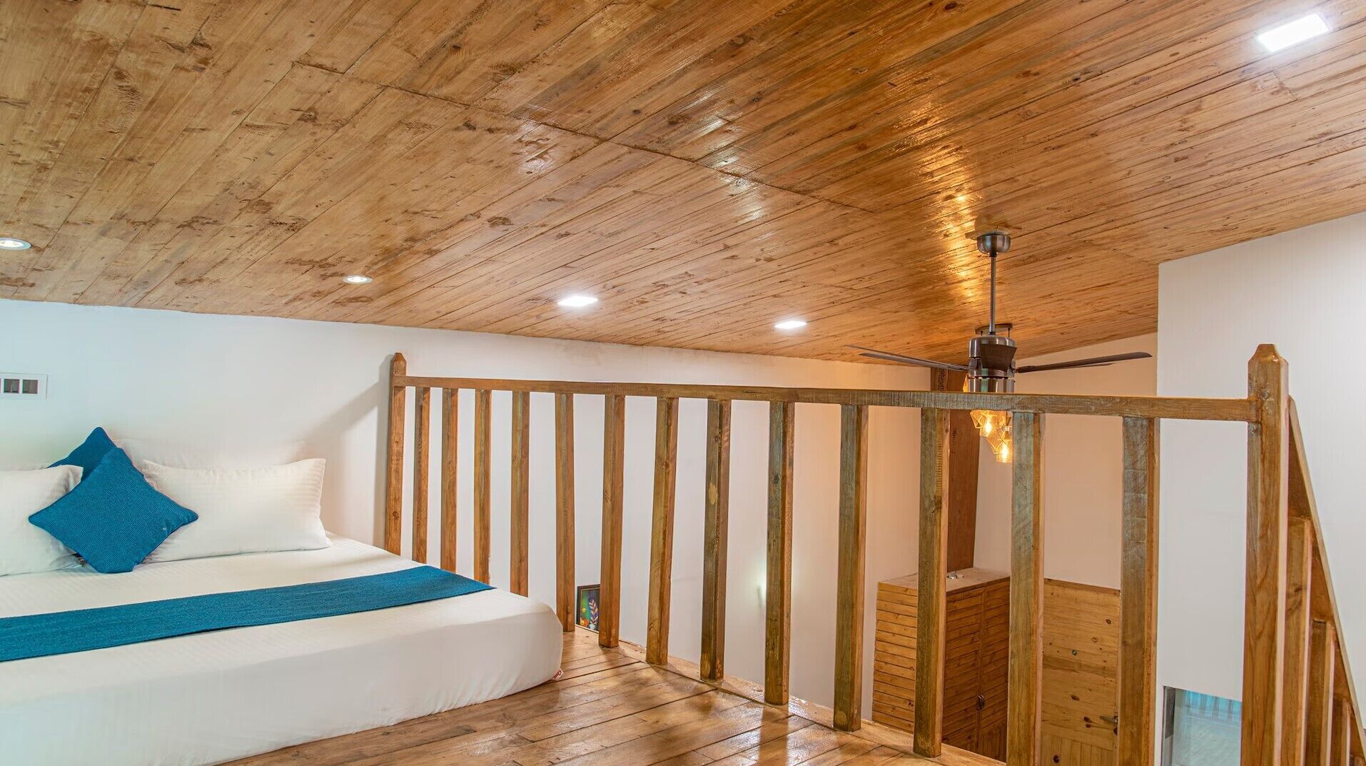 Ein Schlafzimmer mit Holzboden, einem Bett mit weißer Bettwäsche und blauen Kissen, einer Holzreling und einem Deckenventilator.