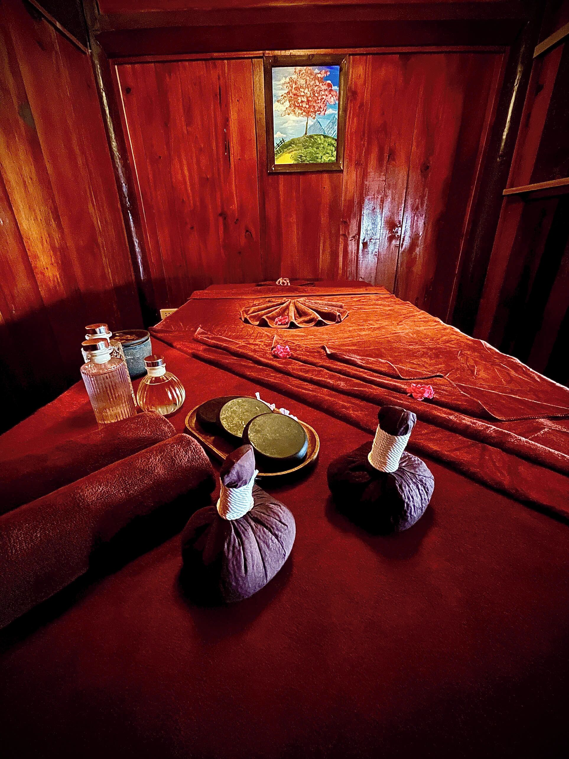 Um quarto com interior em madeira, uma pintura de uma árvore na parede, uma mesa de massagem com toalhas e uma tigela, e uma pequena mesa com garrafas.