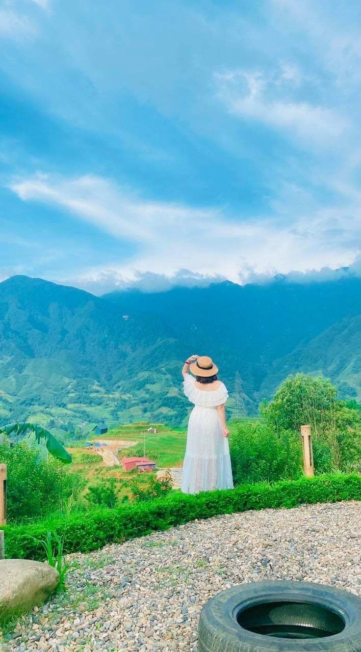 A person in a white dress and hat standing on a gravel path with a view of green hills and a mountain range.