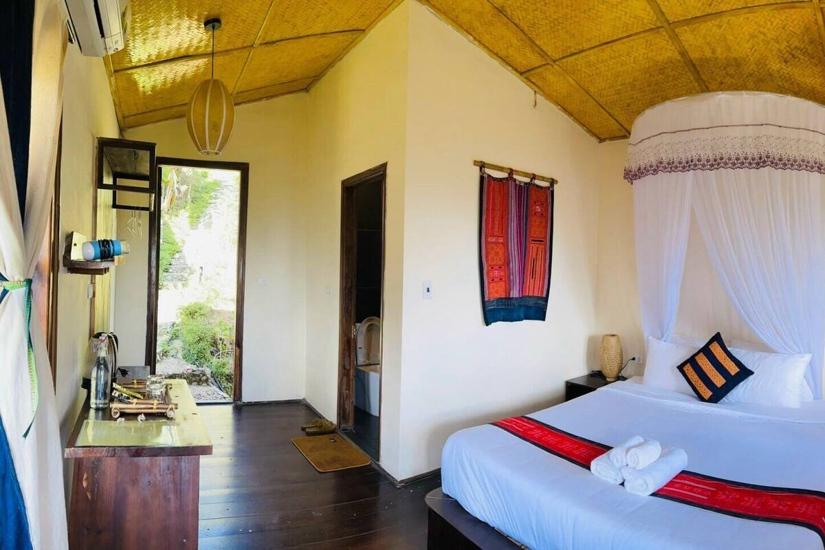 Una habitación luminosa con cama, mesitas de noche, un escritorio pequeño y una ventana con cortinas.