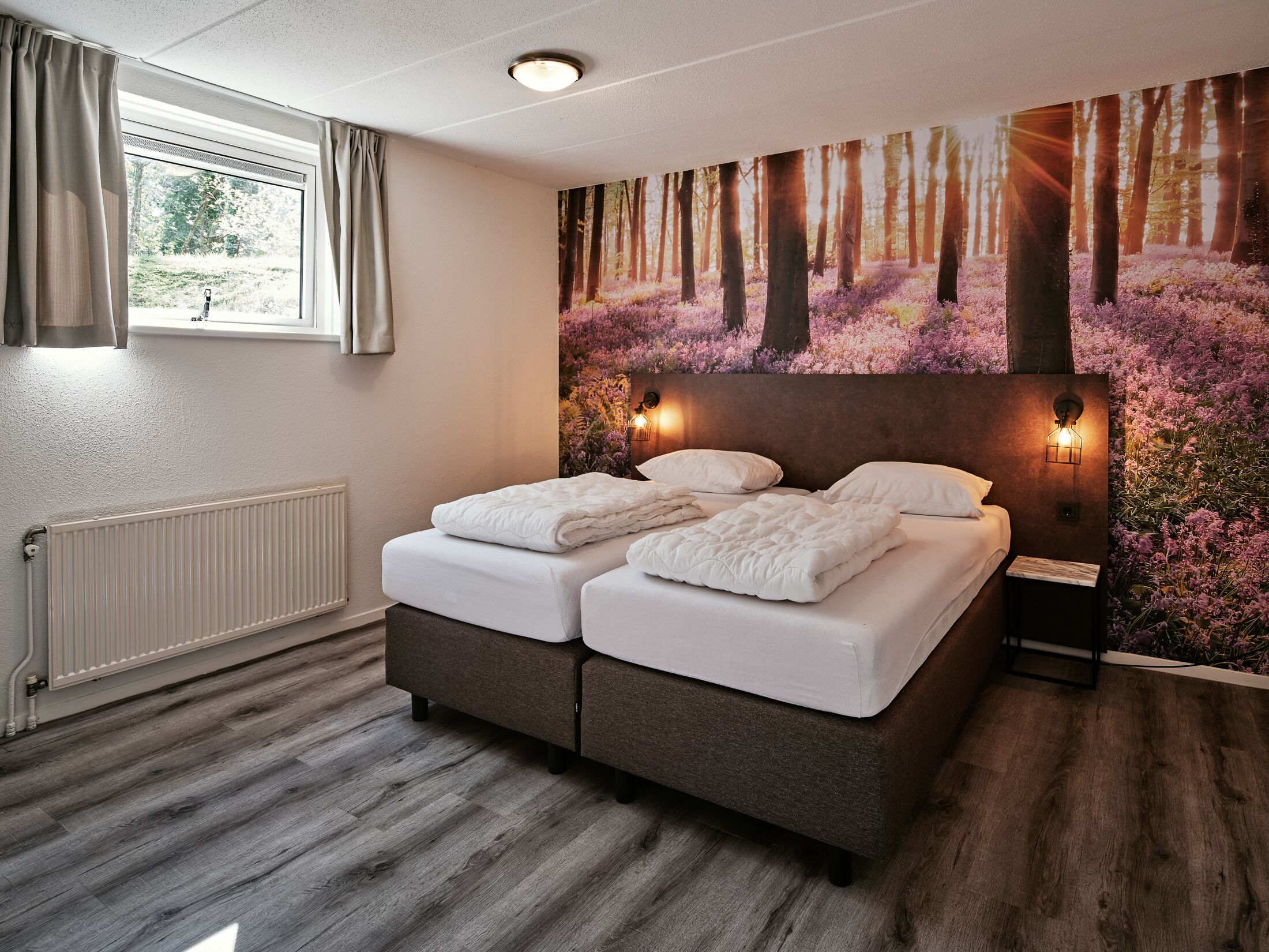 Ein Schlafzimmer mit Doppelbett, Heizkörper, einem Fenster mit Vorhängen und einer Wandmalerei zum Thema Wald.