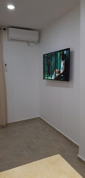 Ett rum med ett kaklat golv, en beige matta, en väggmonterad platt-TV och en luftkonditioneringsanläggning på väggen.