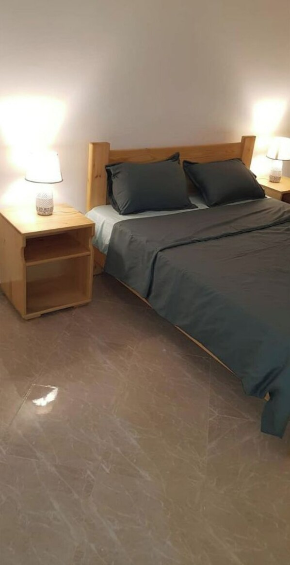 Una cama bien hecha con un edredón oscuro y dos almohadas, flanqueada por mesitas de noche de madera con lámparas.