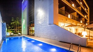 A nighttime swimming pool area with a building featuring a visible neon sign, a staircase, and outdoor seating. - VM Hotel Cumbuco (Caucaia)