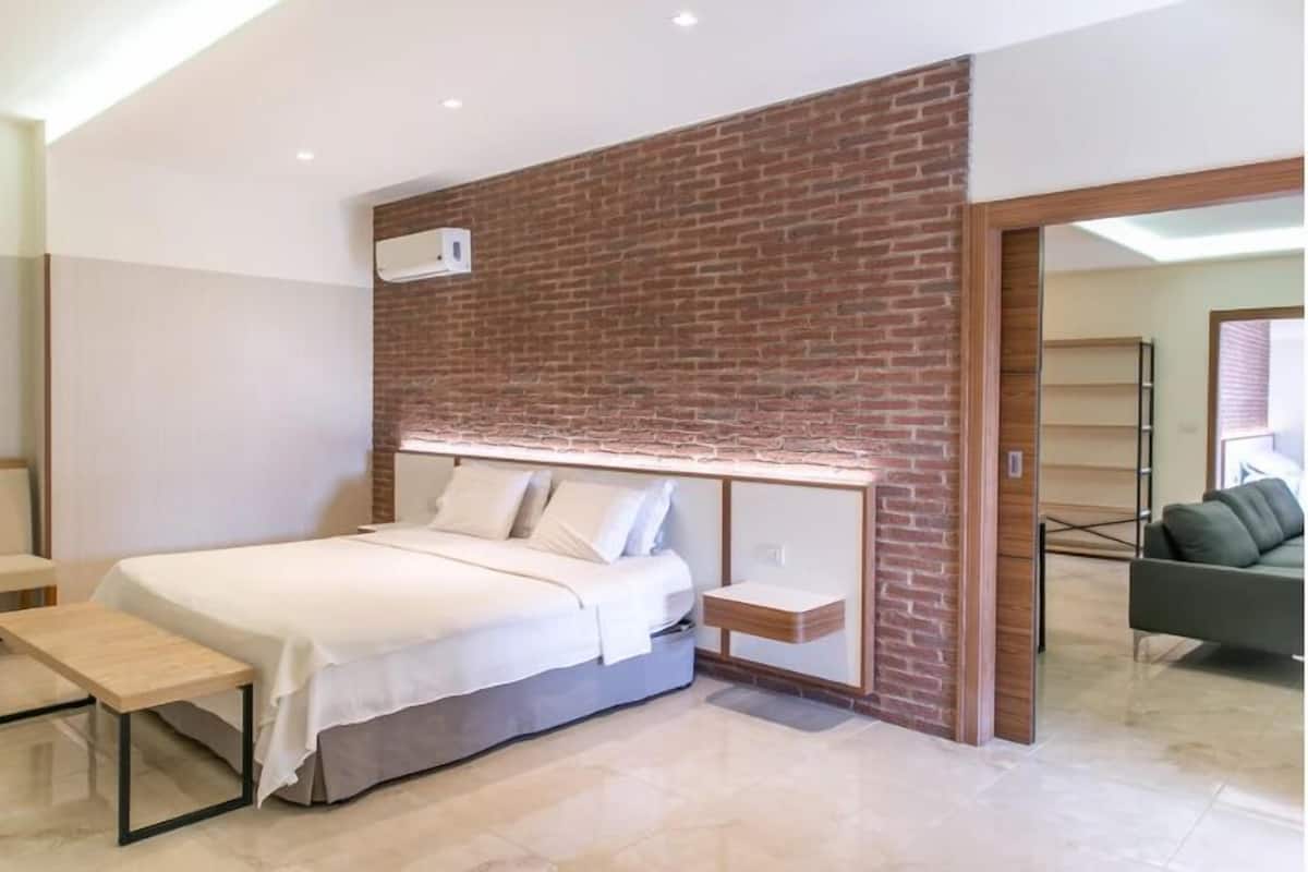 Une chambre moderne avec un mur d’accent en briques, un lit avec du linge de lit blanc, une table de chevet et un canapé vert dans la pièce adjacente.