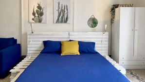 Una camera da letto con una testiera bianca, un letto con lenzuola blu e gialle, una poltrona blu, un armadio e due quadri raffiguranti cactus incorniciati appesi al muro.