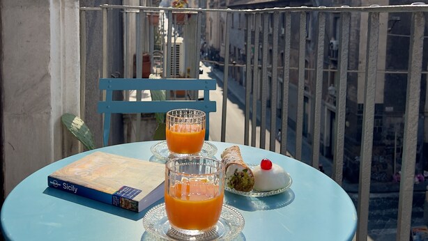 Un balcone con un tavolo apparecchiato per due, con un libro, due bicchieri di succo d'arancia e una pasticceria.