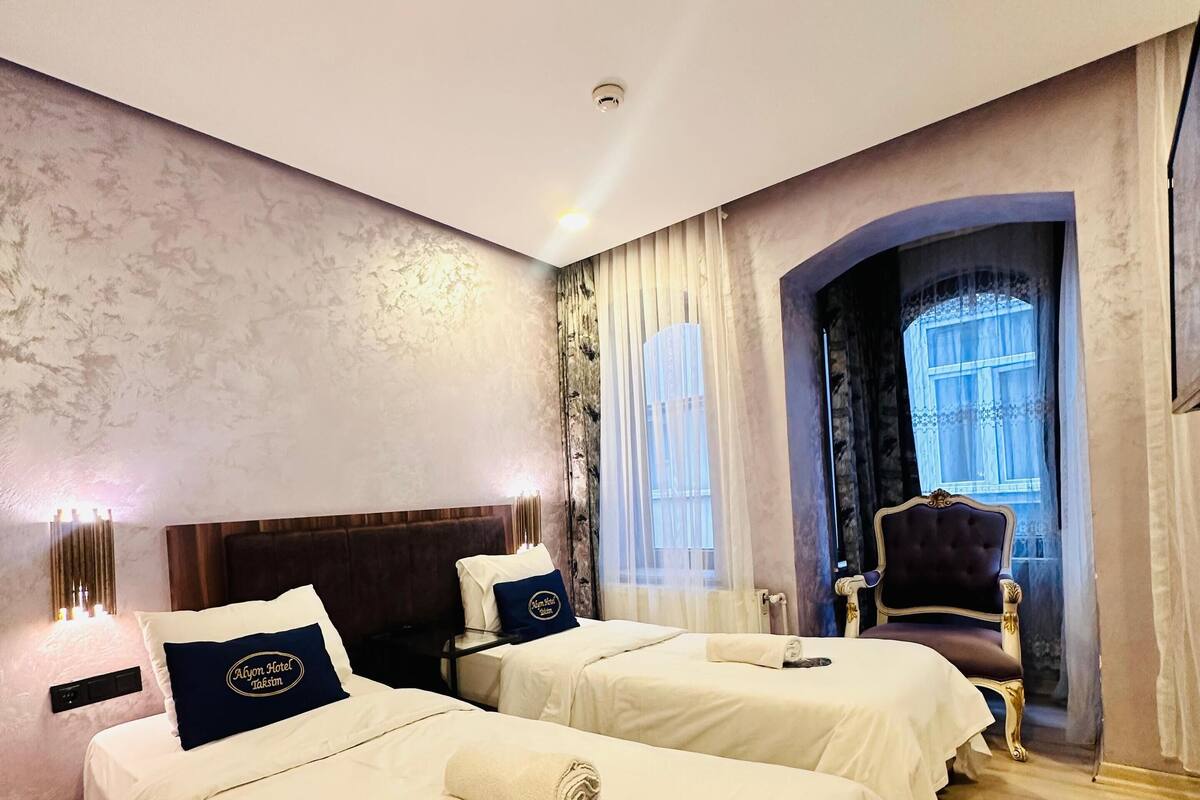 Une chambre d’hôtel avec deux lits, une grande fenêtre avec des rideaux, un plancher en bois et un fauteuil classique.