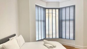 Quarto com uma cama grande, roupa de cama branca e uma toalha enrolada na cama. Há duas janelas com persianas, e o quarto tem piso de madeira e paredes brancas.