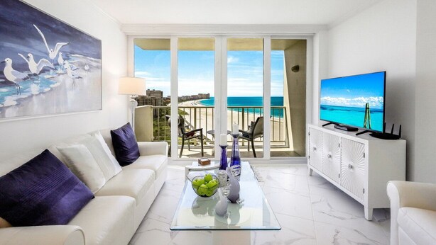 A modern living room with a white sofa, a glass coffee table with decorative items, a flat-screen TV, and a balcony with a view of the sea. - Modern Beachfront Condo | Pool, Gym & Tennis (Marco Island)