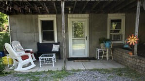 A covered porch with two doors, a seating area with chairs and a table, and a small garden area with a potted plant and decorative items.