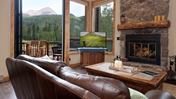 A cozy living room with a stone fireplace, leather sofa, and a television displaying a mountain view. - Corner Cascade Village Condo - Largest deck - Fire Pit - Views - Heated Pool (Durango)