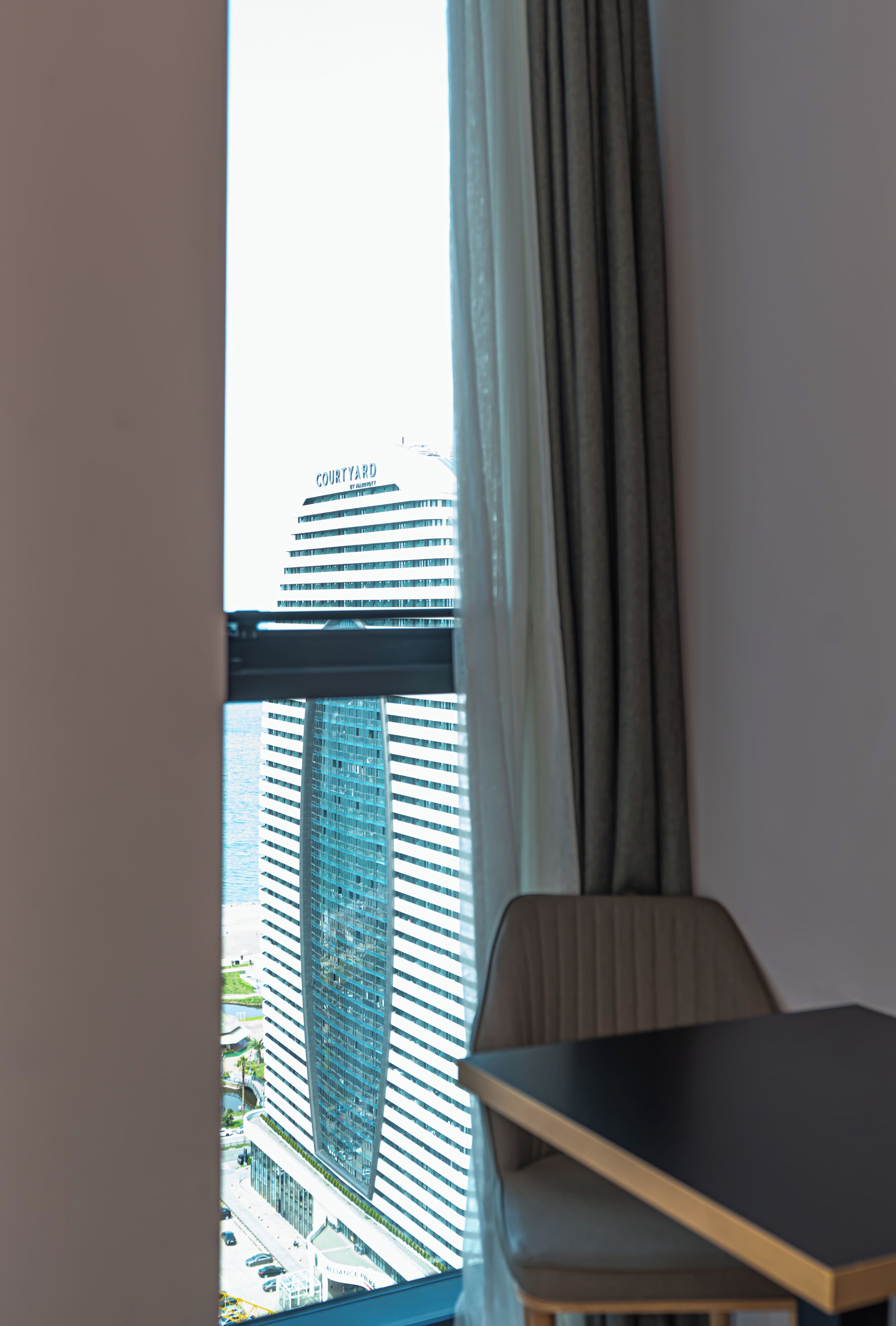 Een kamer met uitzicht op een hoog gebouw, waarop 'COURTYARD' staat, zichtbaar door gordijnen voor het raam.