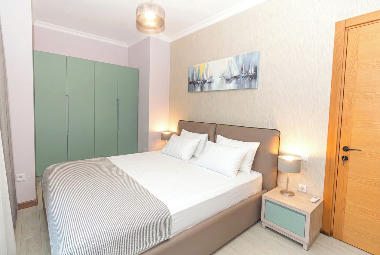 Ein Schlafzimmer mit Bett, Nachttischen, einem grünen Kleiderschrank, einem Gemälde und einer Holztür.