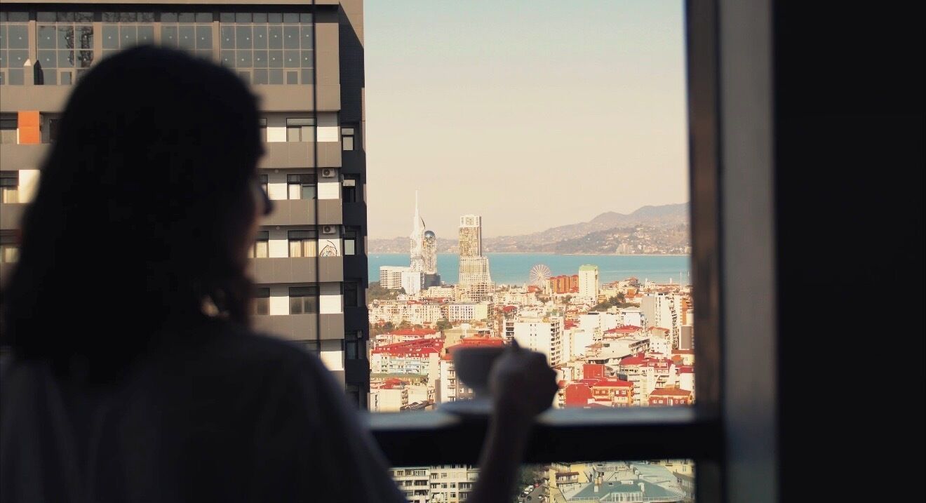 Une personne est assise près d’une fenêtre donnant sur un paysage urbain, avec des bâtiments et une étendue d’eau.