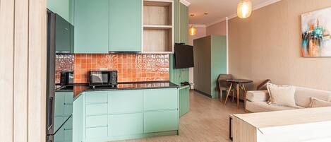 A modern kitchen with teal cabinets, a black countertop, and a microwave. There is a dining area with a table and chairs, and a sofa in the living room.