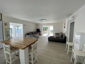 A modern kitchen with a wooden table and white chairs, a grey sofa, and a television in the background.