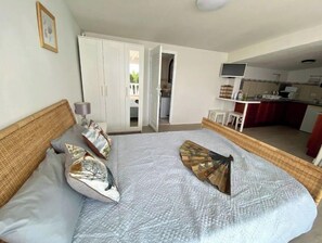 Une chambre à coucher comprenant un lit, des oreillers, une table de chevet, une armoire et une porte ouverte donnant sur un coin cuisine.