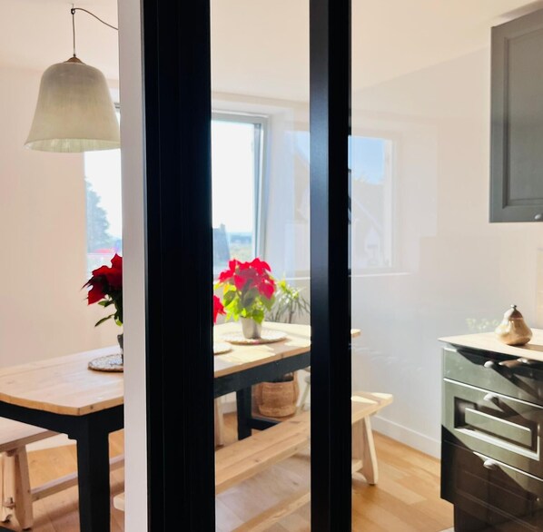 A modern kitchen with a wooden table, a hanging light fixture, and a view of a balcony with red flowers.