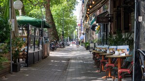 Eine schmale Straße mit Außenbestuhlung und Tischen, einige Fahrräder am Rand geparkt, und ein Baum spendet Schatten.