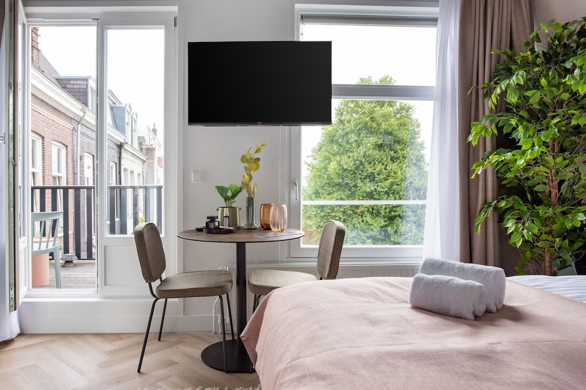 Ein Hotelzimmer mit Bett, Fernseher, Esstisch mit Stühlen, einer Zimmerpflanze und Blick auf Gebäude und Bäume.