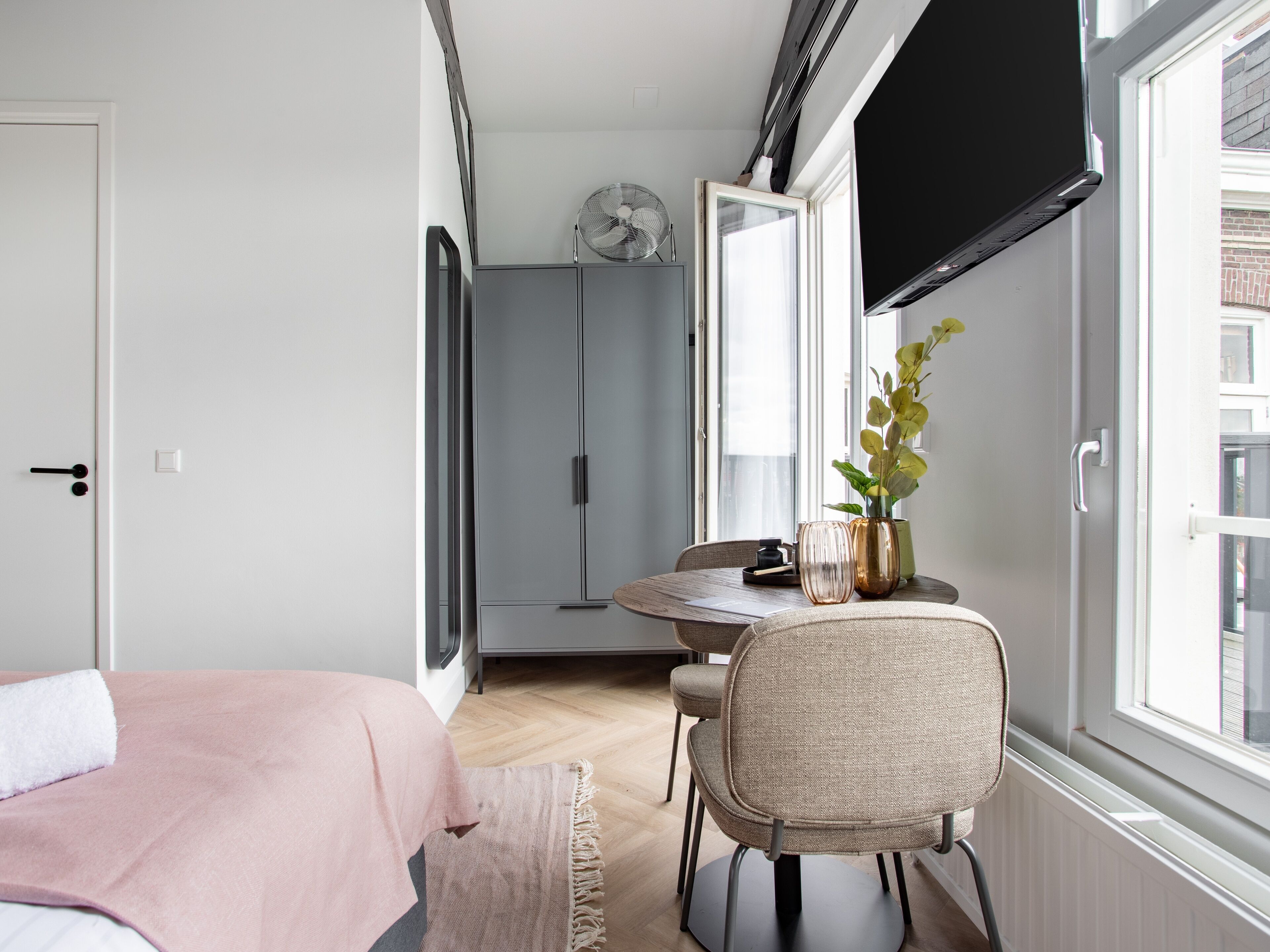Een compacte kamer met een bed, een ronde eettafel met stoelen, een klein tafeltje met een vaas en een ventilator, en een aan de muur bevestigde televisie.