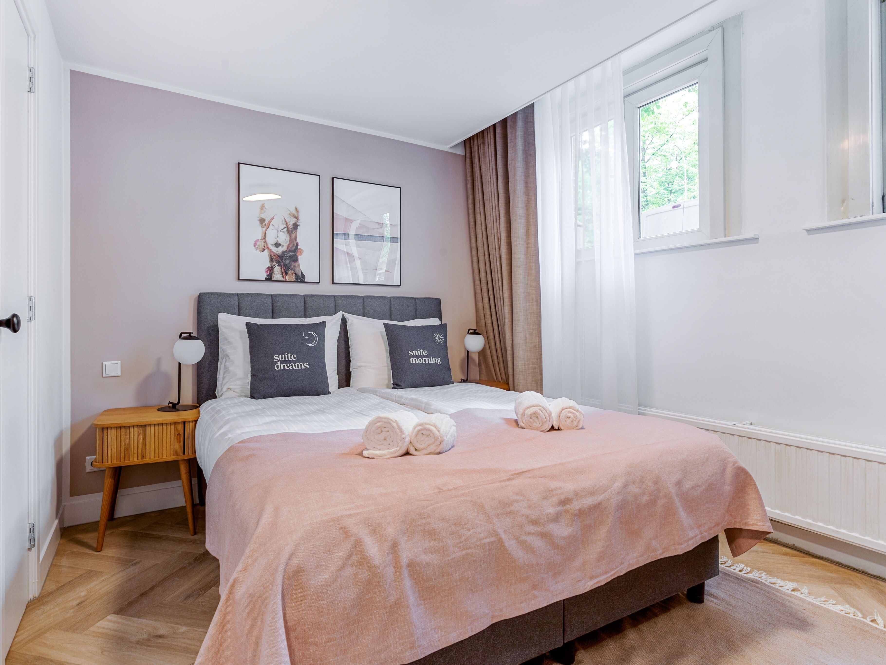 Un dormitorio con una cama, dos mesitas de noche, un velador, un ventanal con cortinas y dos cuadros colgados en la pared.