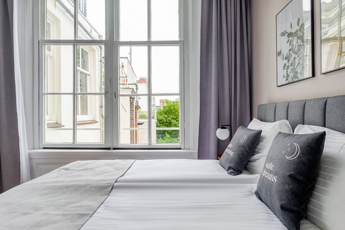 Ein Hotelzimmer mit großem Fenster, einem Bett mit weißen Leinen, einem grauen Kopfteil und zwei Zierkissen.
