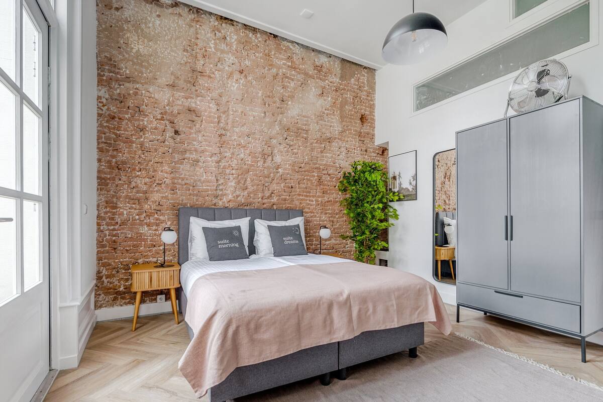 Ein modernes Schlafzimmer mit Backsteinwand, grauem Bett, großem Kleiderschrank und einer Topfpflanze.