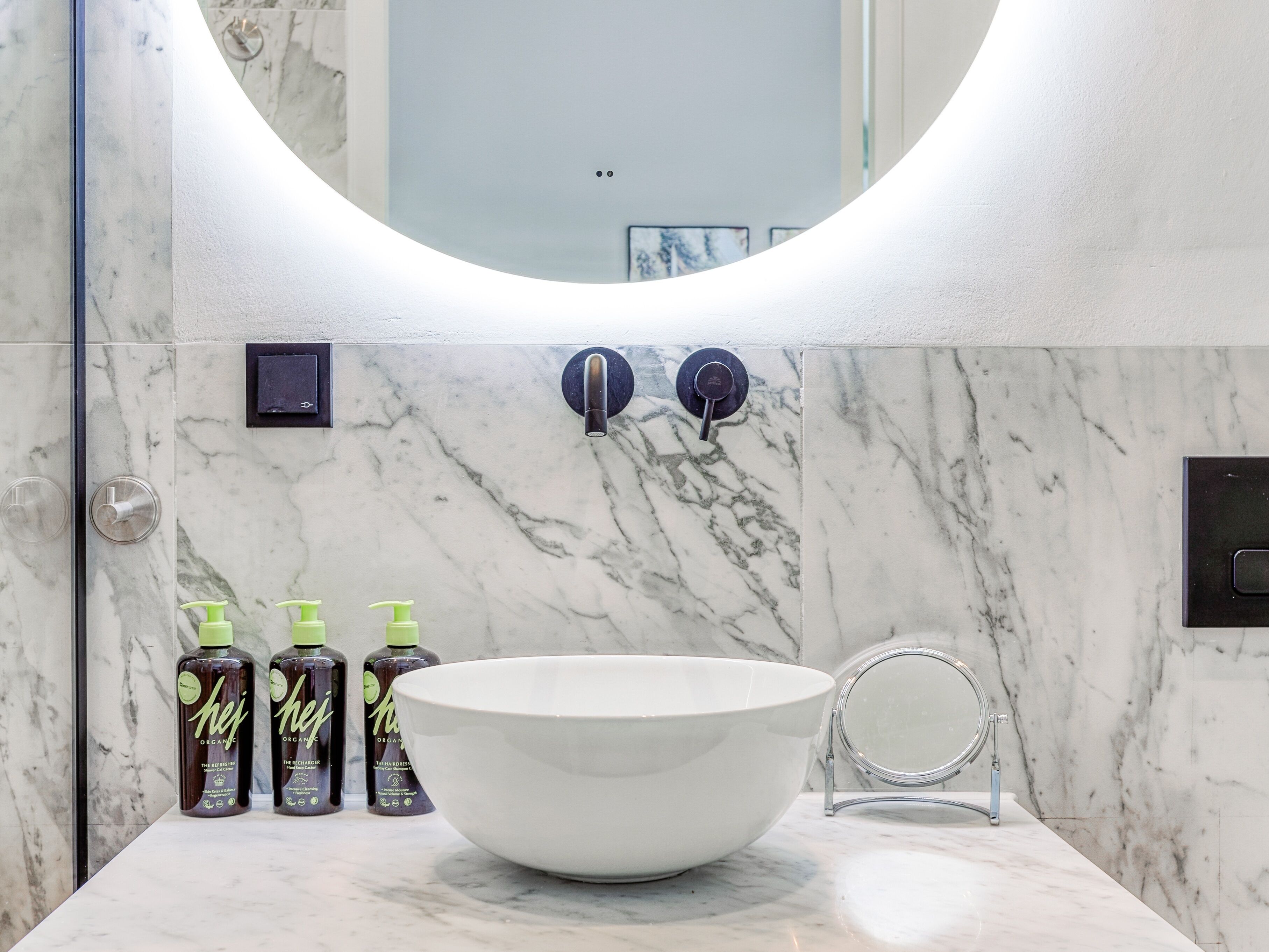 Une salle de bain avec un lavabo en marbre blanc, un grand miroir et trois bouteilles de produits capillaires biologiques.
