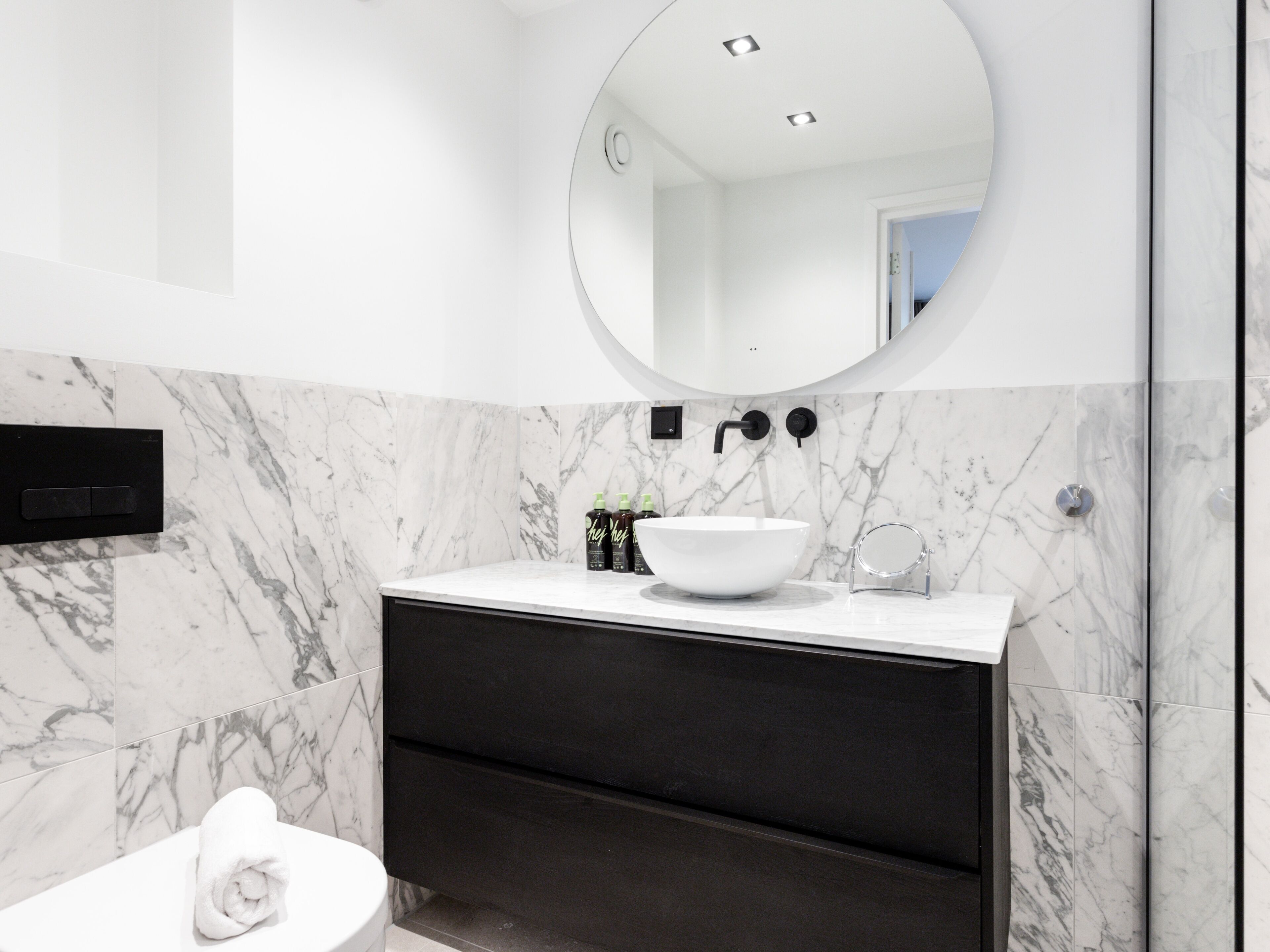 Een moderne badkamer met een wit marmeren wastafel, een donkere badkamermeubel en een ronde spiegel.