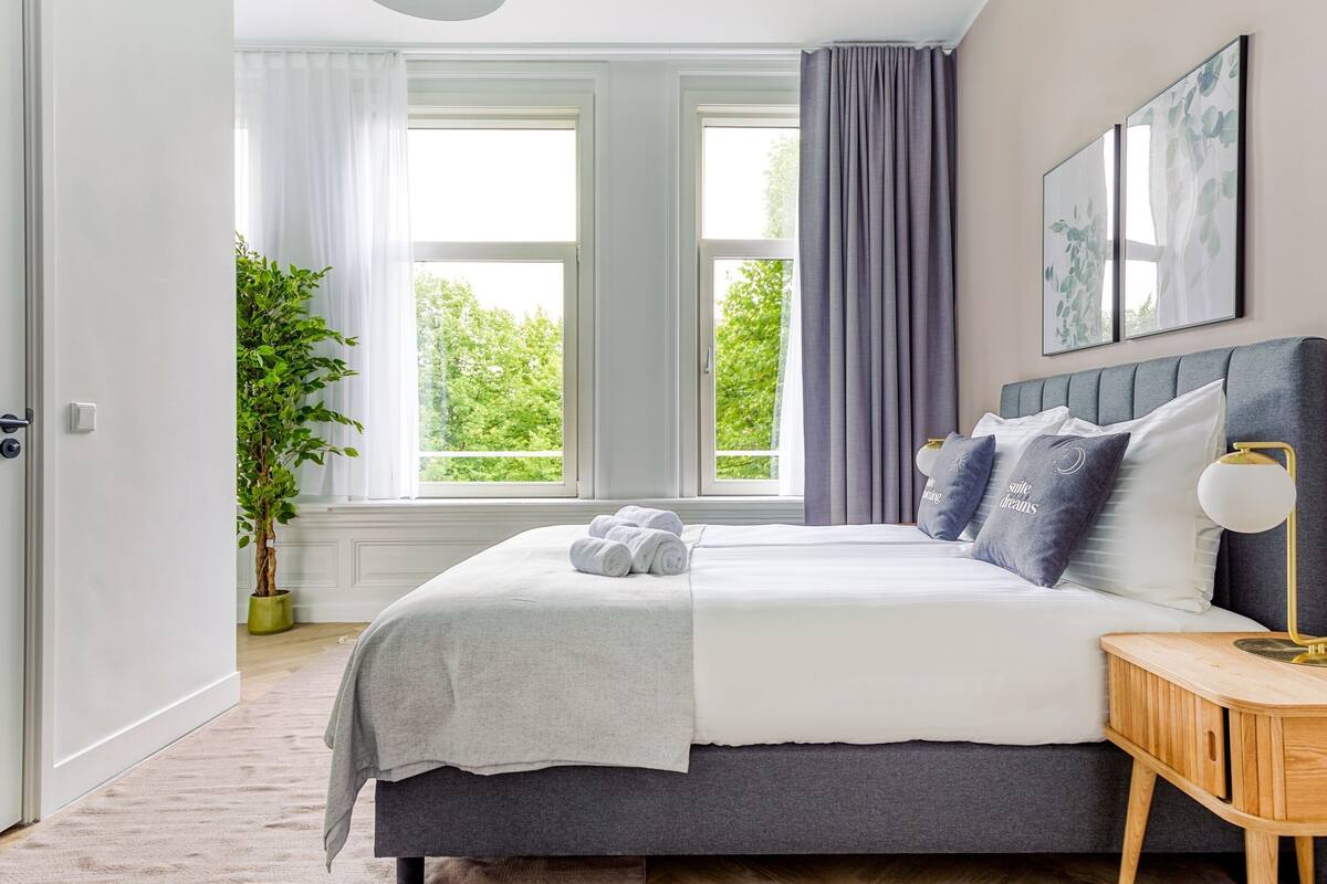 Ein Schlafzimmer mit Bett, Nachttisch, Lampe, Fenster und einer Pflanze.