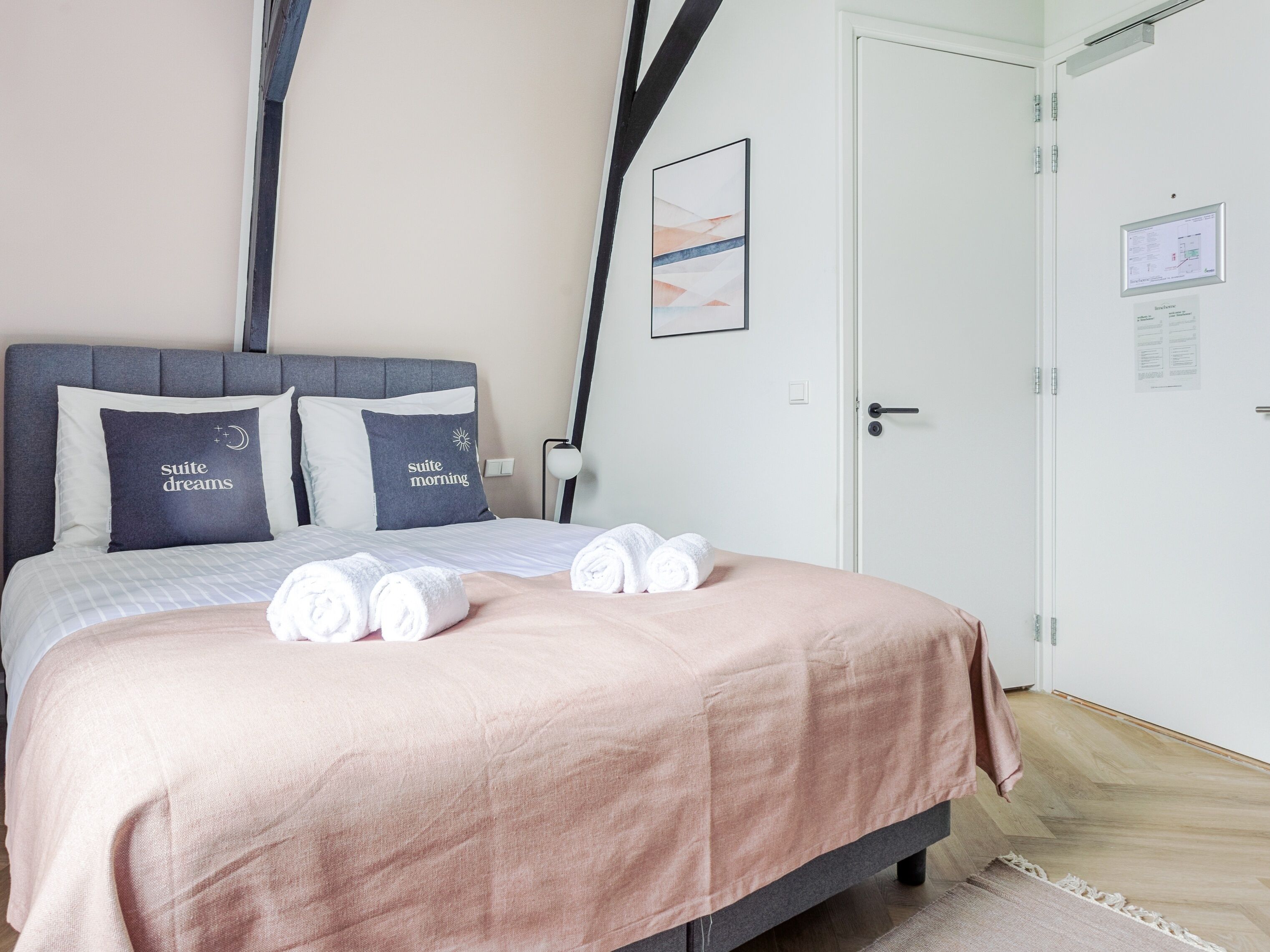 A neatly made bed with a pink bedspread, two pillows with text, and rolled towels on the bed.