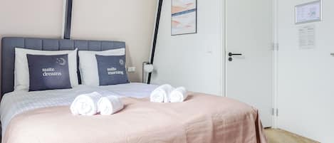 A neatly made bed with a pink bedspread, two pillows with text, and rolled towels on the bed.