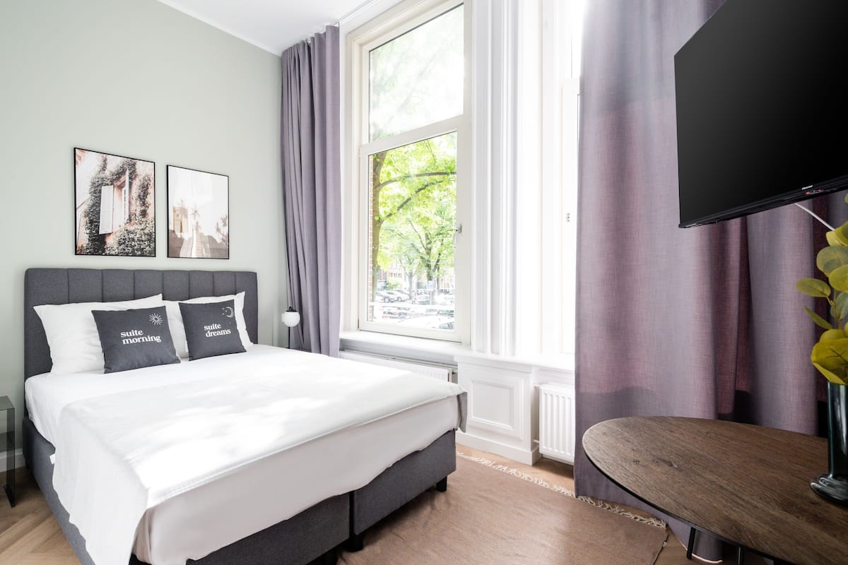 Ein modernes Hotelzimmer mit einem großen Bett, einem Flachbildfernseher, einem Fenster mit Vorhängen und einem Kunstwerk an der Wand.