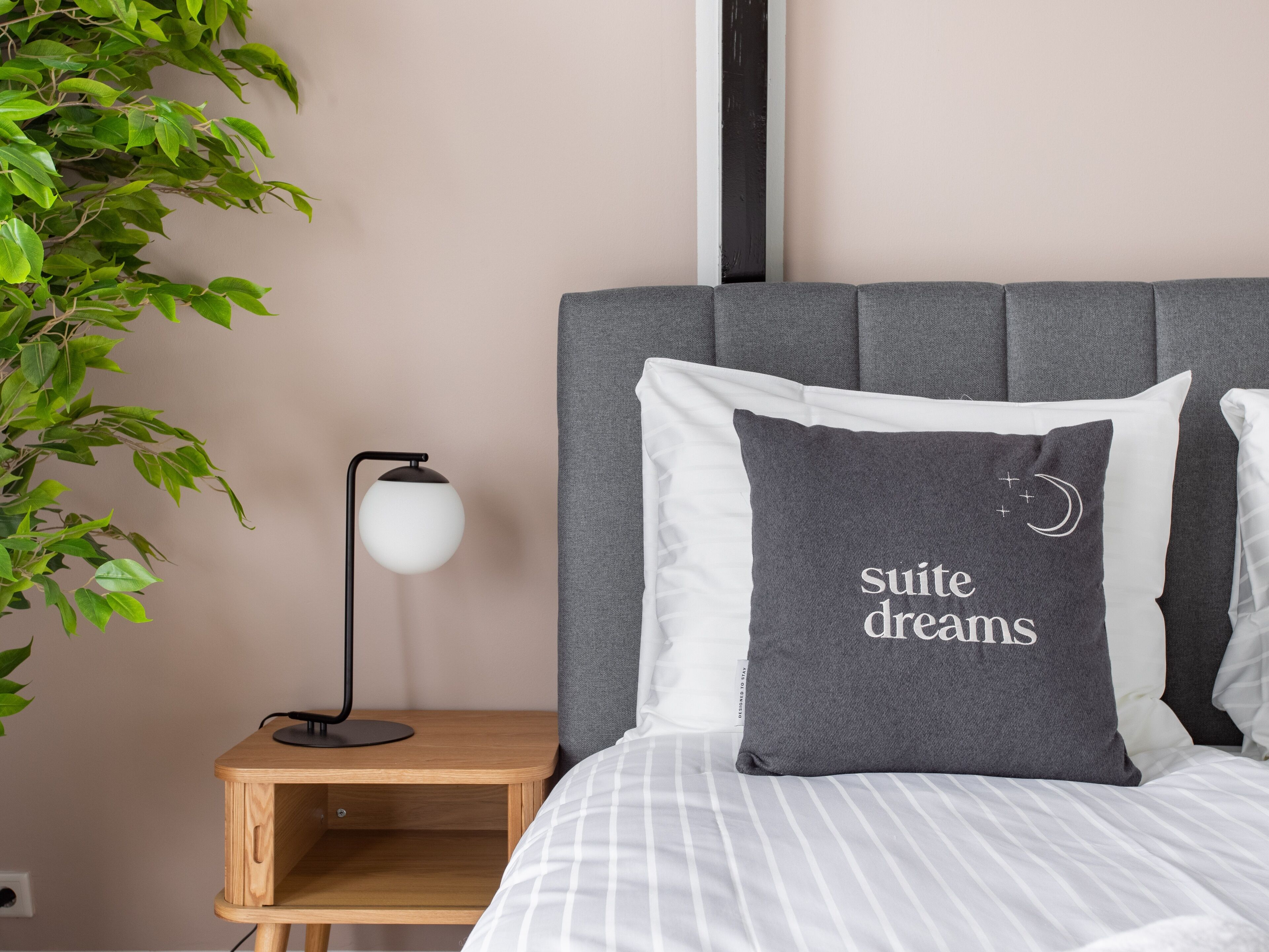 Una cama impecablemente arreglada con ropa de cama a rayas, un almohadón gris con el texto "suite dreams" y una lámpara de noche, además de una mesita de noche de madera.