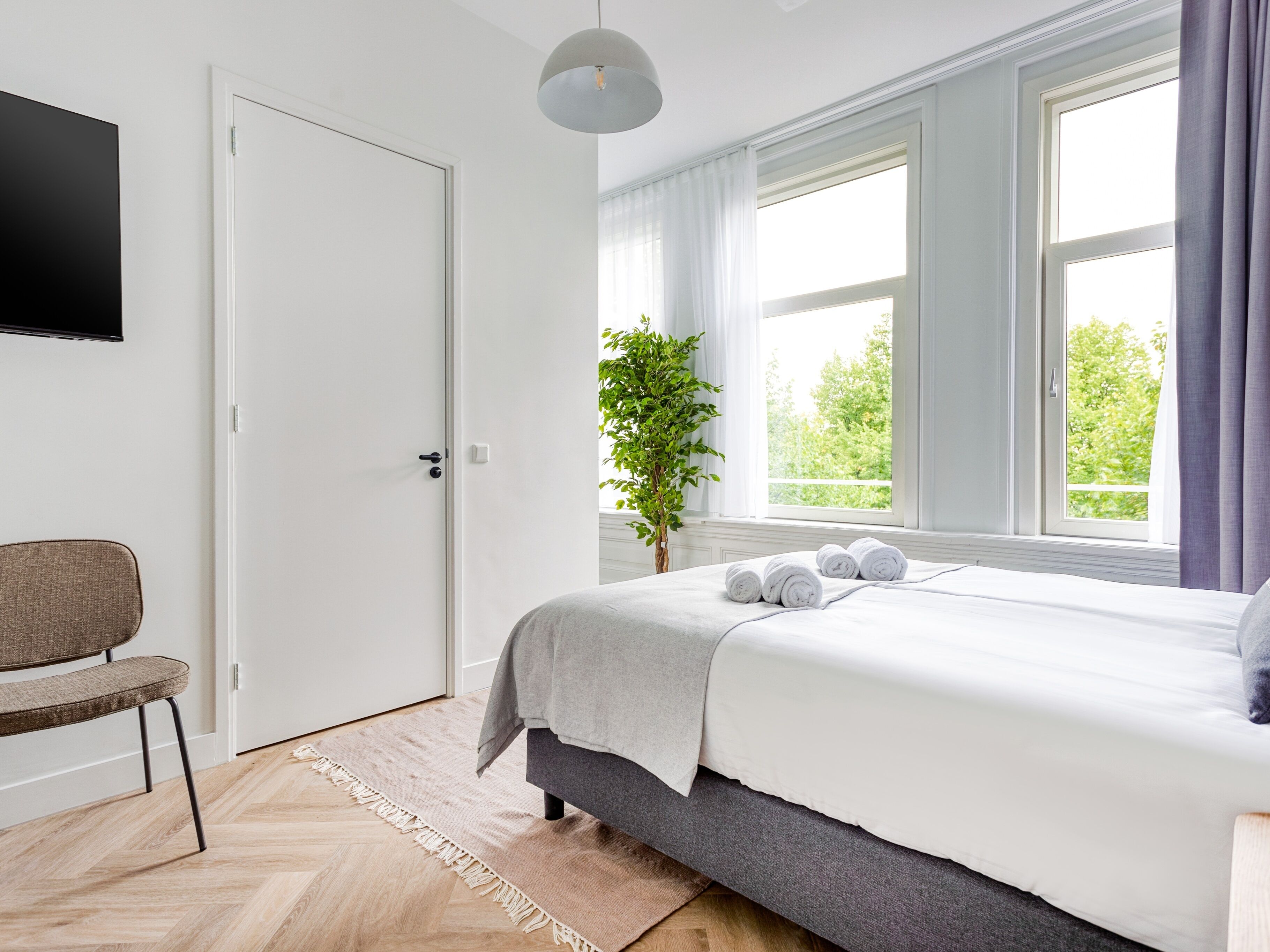Quarto com cama, uma cadeira, uma planta e televisão na parede.