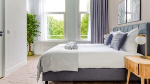 Ein modernes Schlafzimmer mit einem großen Bett, einem Nachttisch, einer Zimmerpflanze und einem Fenster mit Vorhängen.