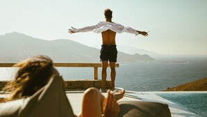 Iemand staat op een balkon, armen wijd, met uitzicht op een wateroppervlak en bergketens in de verte.