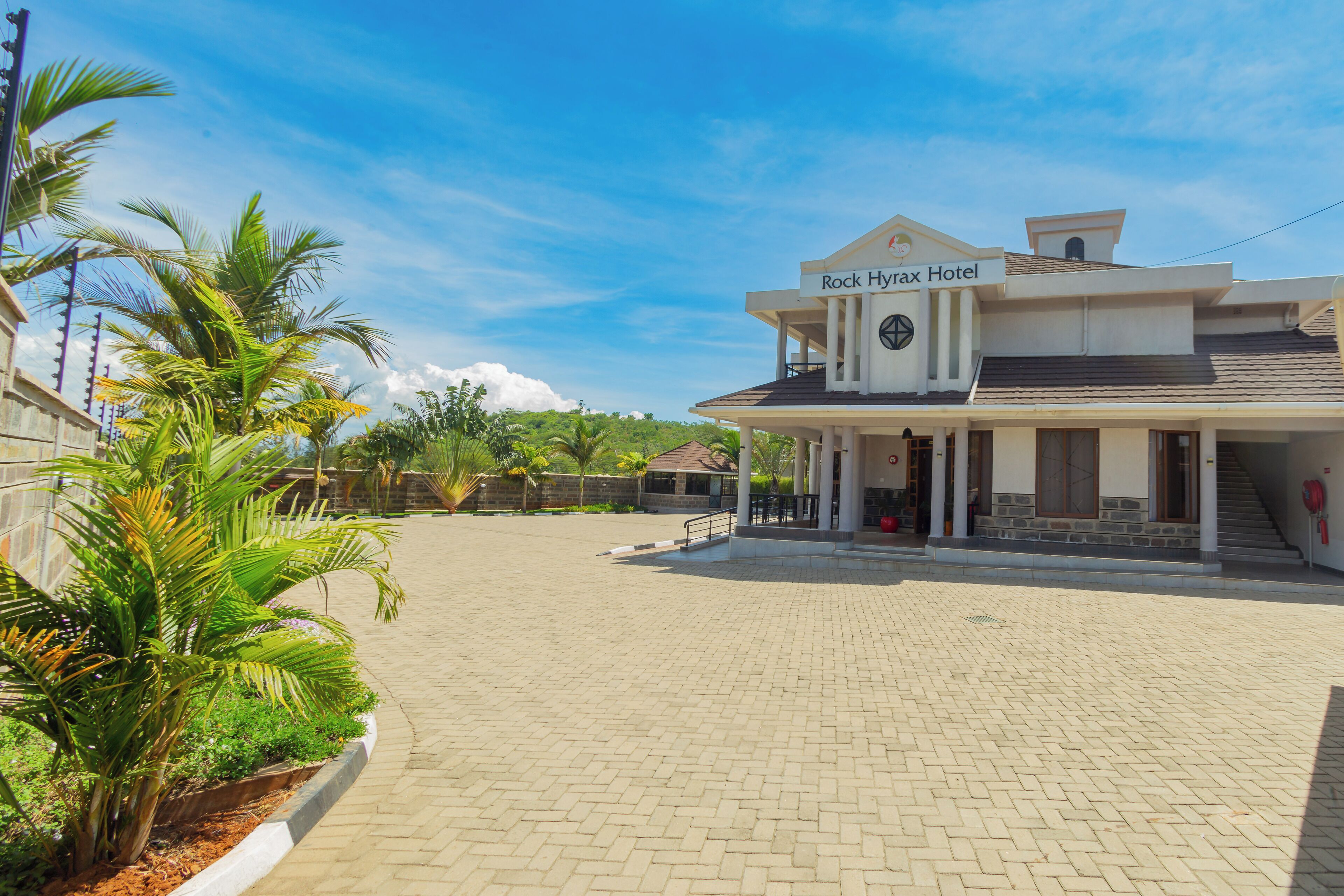 A hotel with a paved courtyard, palm trees, and a sign reading "Rock Hyrax Hotel".