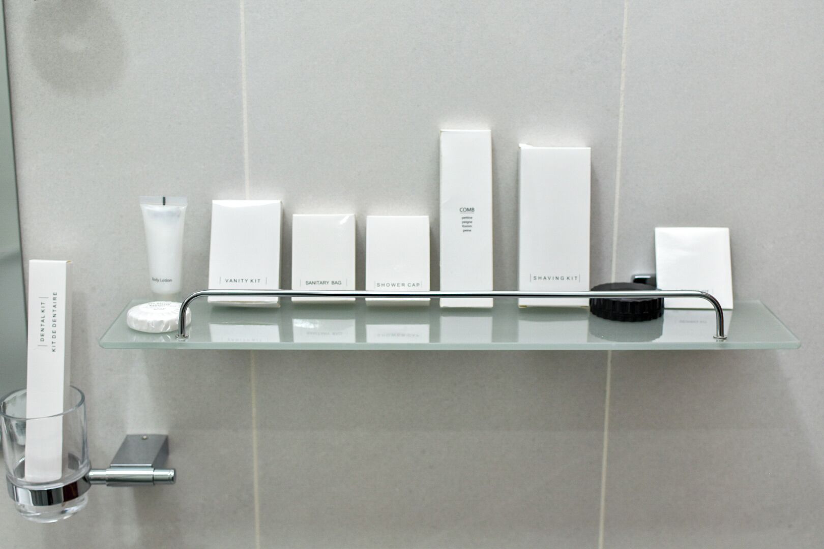 A bathroom with a glass shelf holding various toiletries including a dental kit, vanity kit, sanitary bag, shower cap, comb, shaving kit, and body lotion.