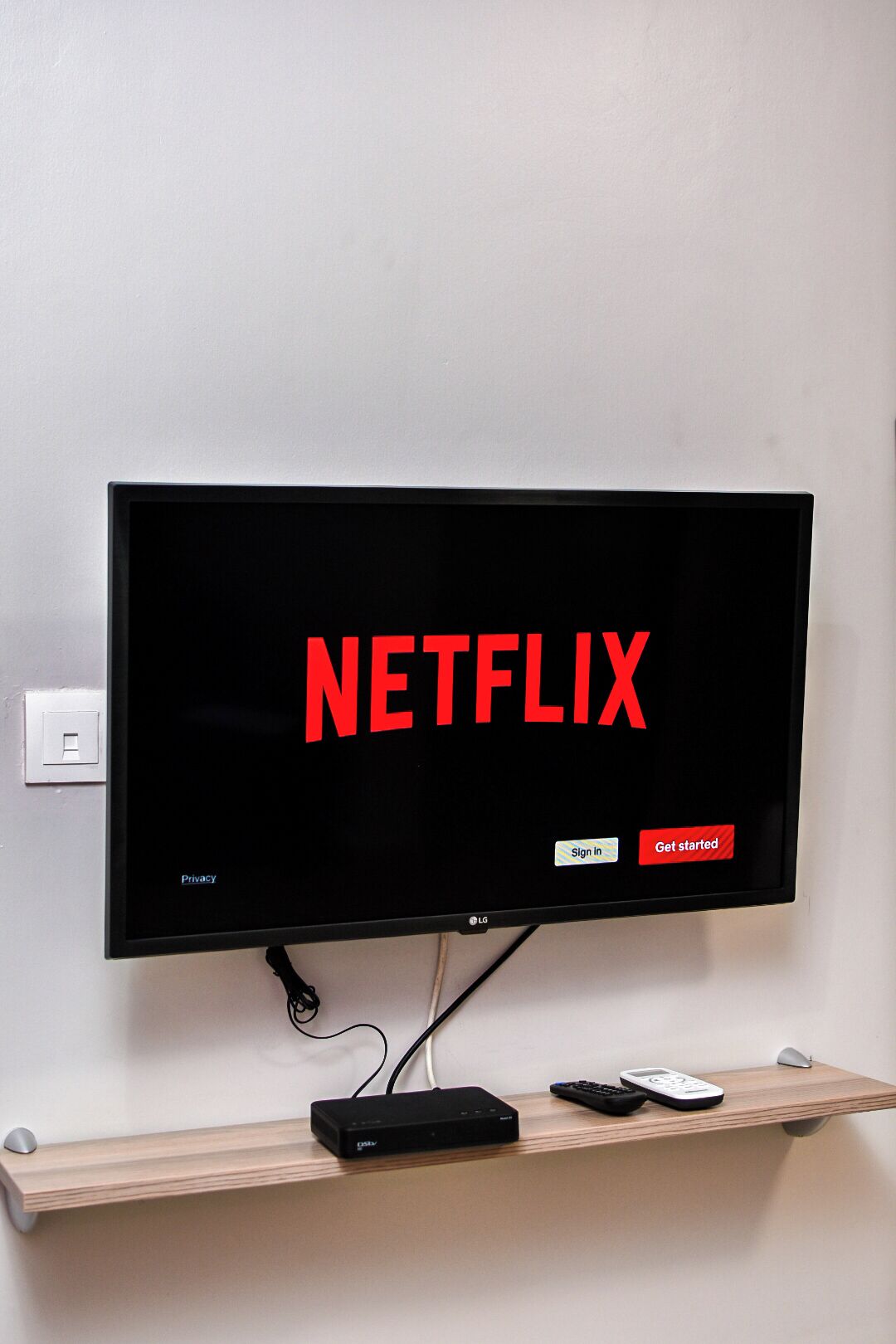 A flat-screen TV mounted on a wall with a wooden shelf below displaying a streaming device and a remote control.