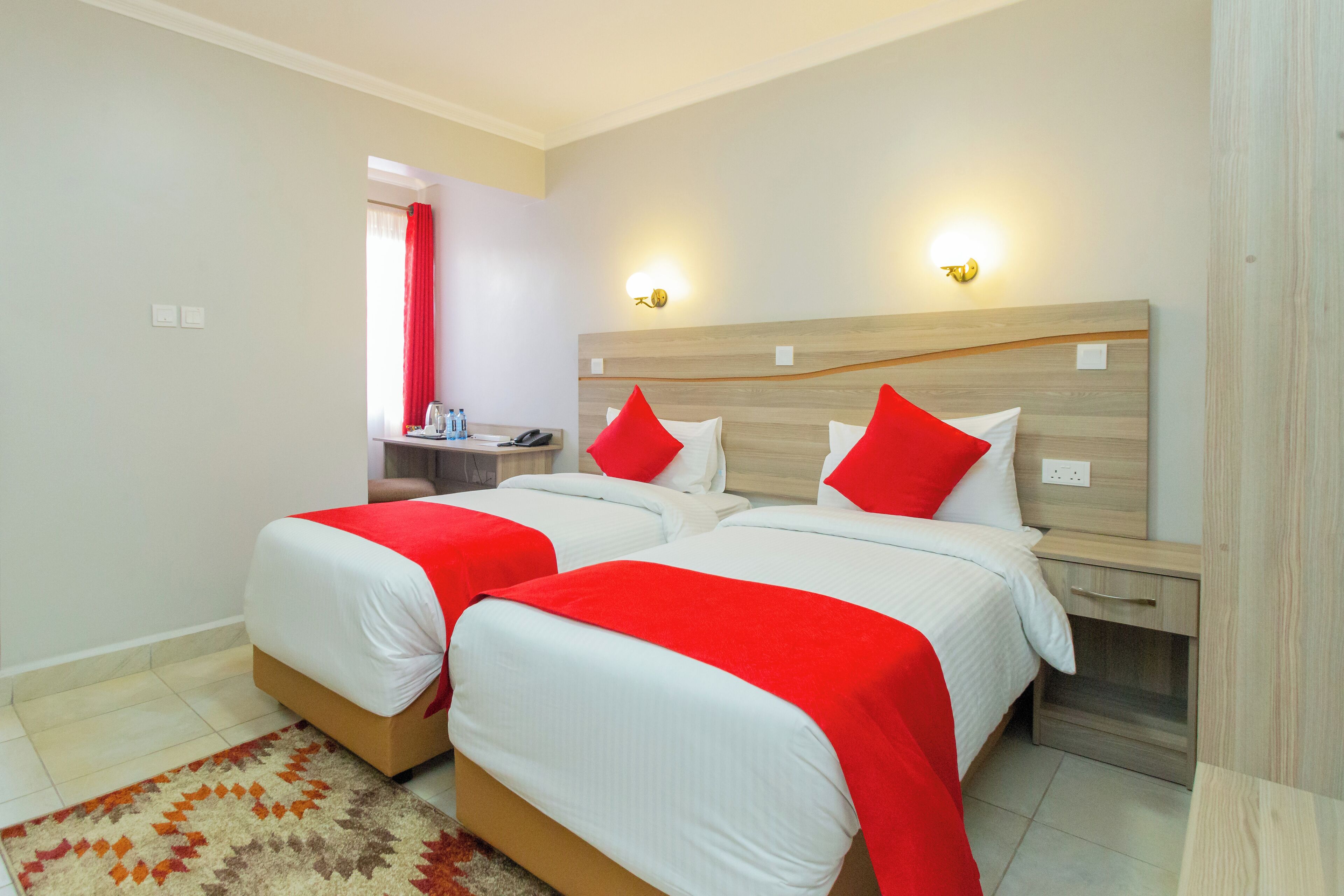 A hotel room with two beds, each with red and white bedding, a wooden headboard, bedside tables, and a small desk with a chair.