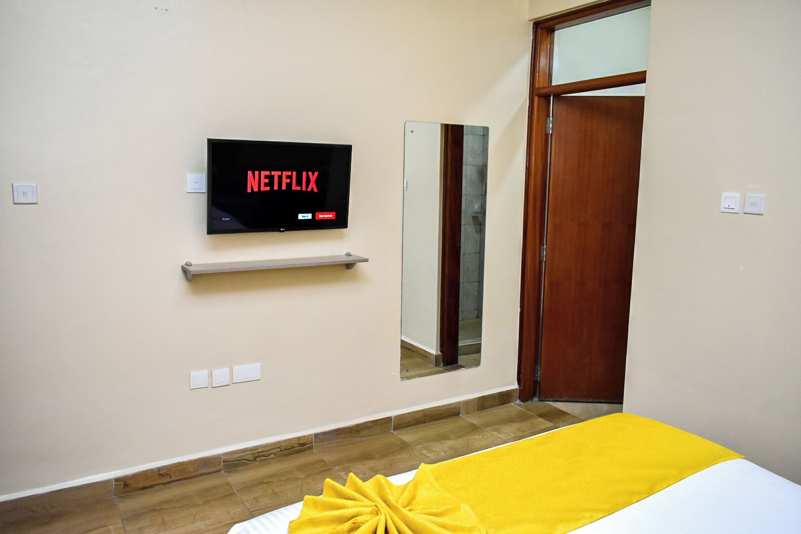 A hotel room with a television mounted on the wall displaying the Netflix logo, a mirror, and a wooden door.