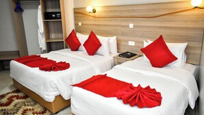 A hotel room with two beds, each with white bedding and red pillows. There is a wooden headboard, a nightstand with a book, and a wardrobe with a blue hanger. - Rock Hyrax Hotel (Nakuru)