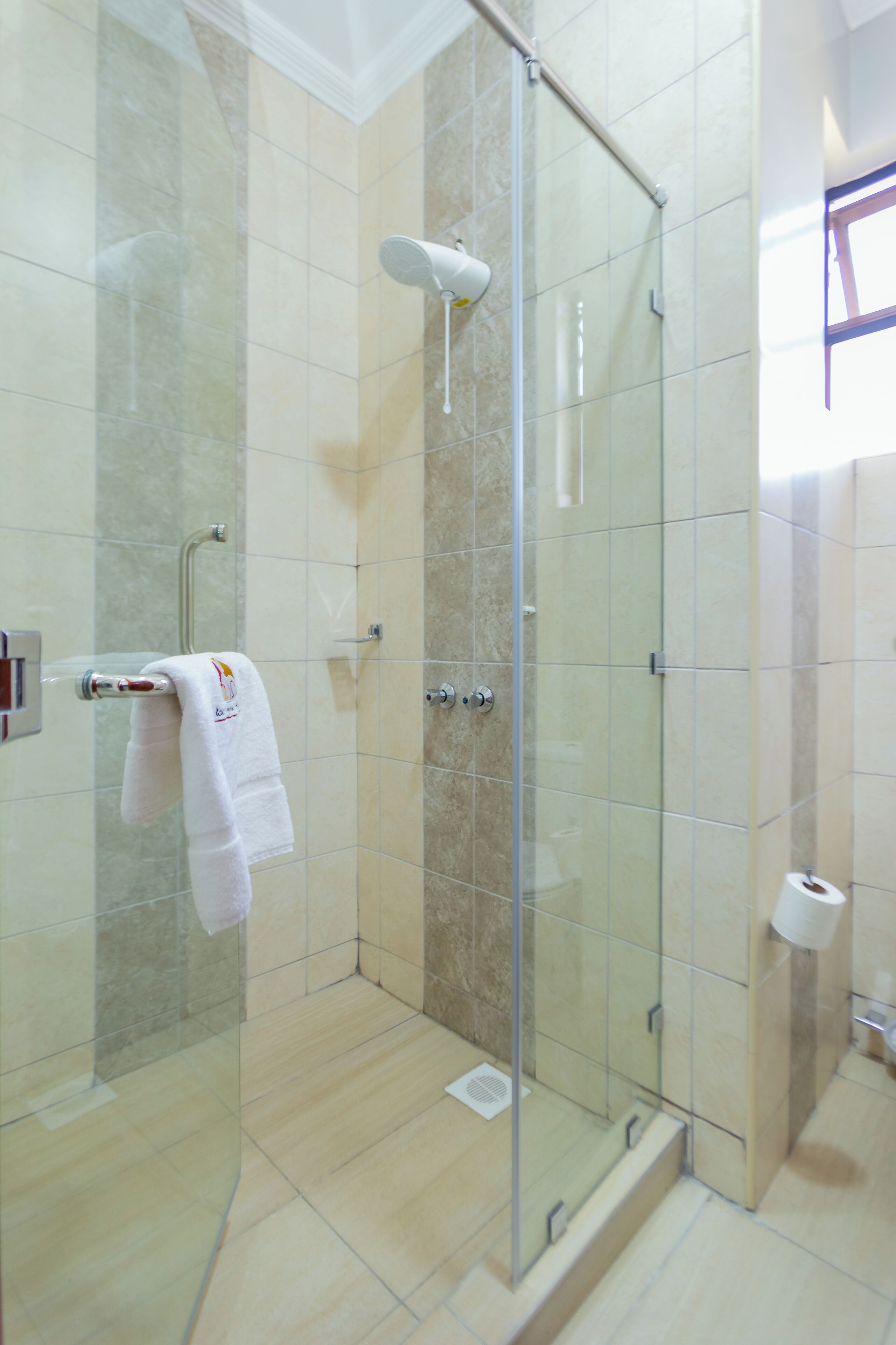 Eine begehbare, gläserne Duschkabine mit einem Handtuch auf einem Halter, einer Handbrause und einem Toilettenpapierhalter an der Wand.