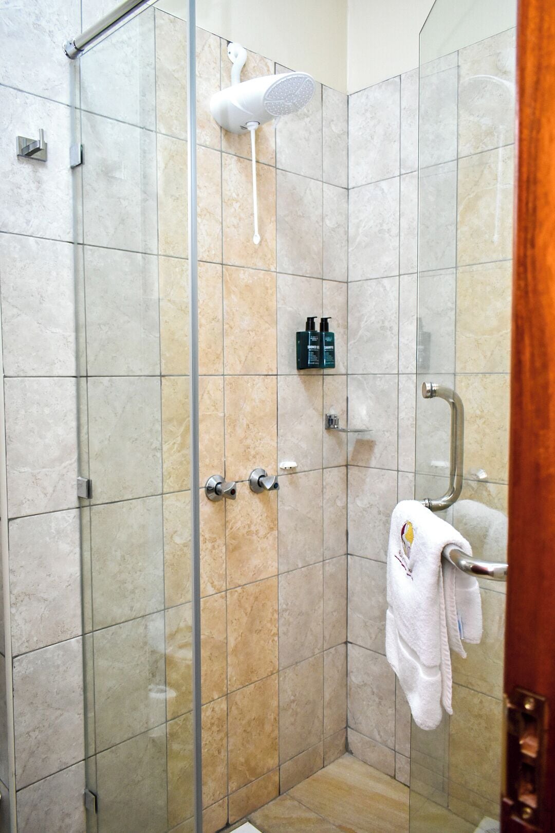 A glass-enclosed shower with a handheld showerhead, a towel rack with a towel, and a shelf with toiletries.
