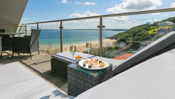 A balcony with a view of the beach and ocean, wicker furniture, and a glass table with food and drinks. - Porthminster Penthouse (St Ives)