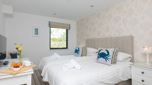 A hotel room with two beds, a desk with a laptop, a vase with flowers, and a window with a view of greenery. - Porthminster Penthouse (St Ives)