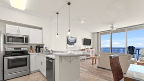 Une cuisine moderne avec des meubles blancs, des appareils en acier inoxydable et un îlot central. On y trouve un coin repas avec un canapé et un téléviseur, ainsi qu’un balcon avec vue sur la mer.